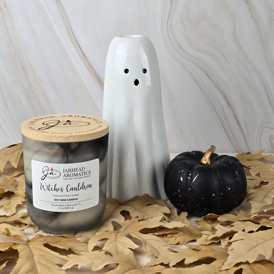 A white ceramic ghost figurine, a black pumpkin, and a Jarhead Aromatics Witches Cauldron - Soy Wax Candle from the Halloween Collection sit on a table with tan leaves, set against a marble-patterned background.