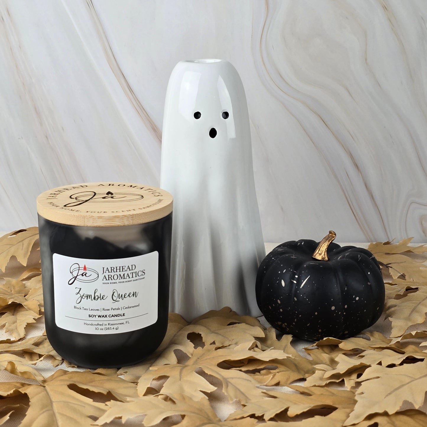 A black Zombie Queen - Soy Wax Candle from Jarhead Aromatics Halloween Collection sits with a white ceramic ghost and small black pumpkin on dried leaves against a light marble background.