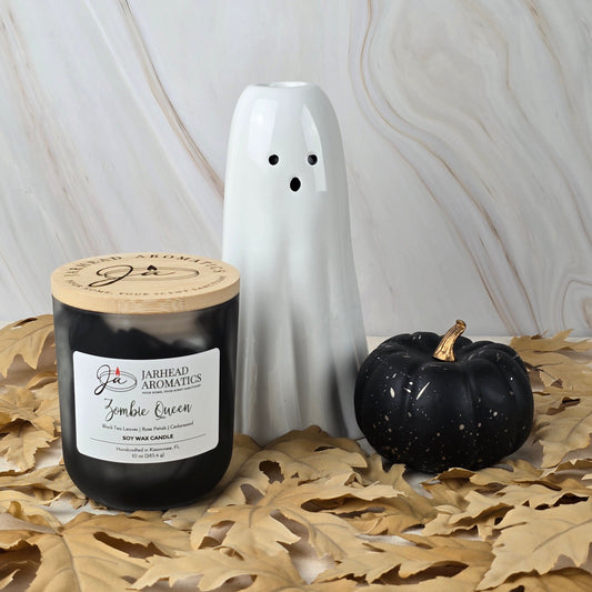 A black Zombie Queen - Soy Wax Candle from Jarhead Aromatics Halloween Collection sits with a white ceramic ghost and small black pumpkin on dried leaves against a light marble background.