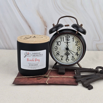 A black Beach Day - Soy Wax Candle by Jarhead Aromatics, featuring a fresh summer scent, sits near a vintage black alarm clock set to 8:00, old keys, and a leather-bound book on beige against a marbled backdrop.