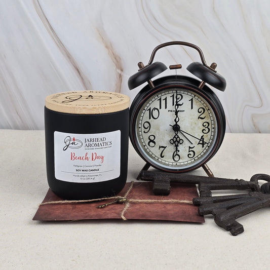 A black Beach Day - Soy Wax Candle by Jarhead Aromatics, featuring a fresh summer scent, sits near a vintage black alarm clock set to 8:00, old keys, and a leather-bound book on beige against a marbled backdrop.