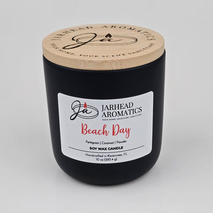 A black jar candle with a wooden lid from Jarhead Aromatics, labeled Beach Day Soy Wax Candle | Coconut, Citrus & Coastal Breeze Candle. This handcrafted candle from Kissimmee, FL offers a long-lasting coastal fragrance blend of coconut and citrus notes with hints of coastal breeze.