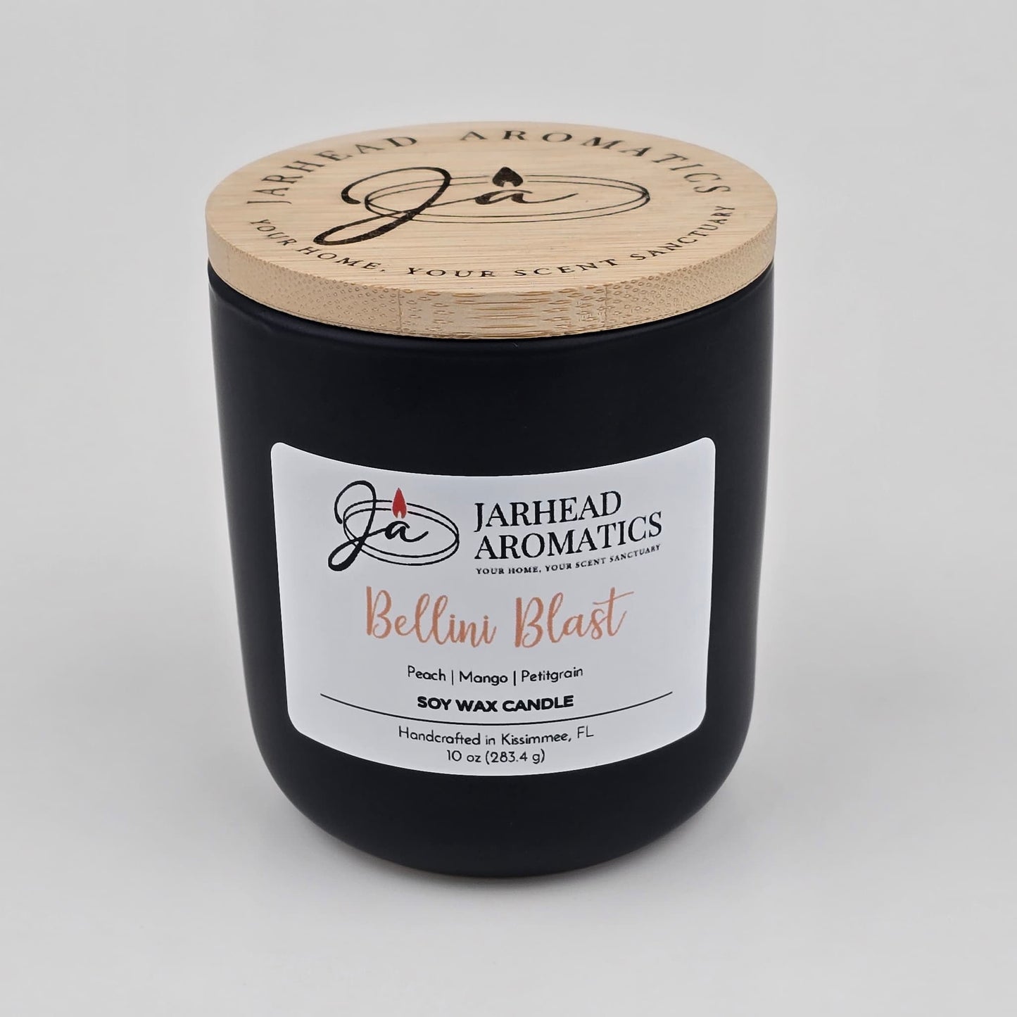 A black candle jar with a wooden lid labeled Jarhead Aromatics. The Bellini Blast Soy Wax Candle features peach, mango, and tangerine scents. 10 oz, handcrafted in Kissimmee, FL.