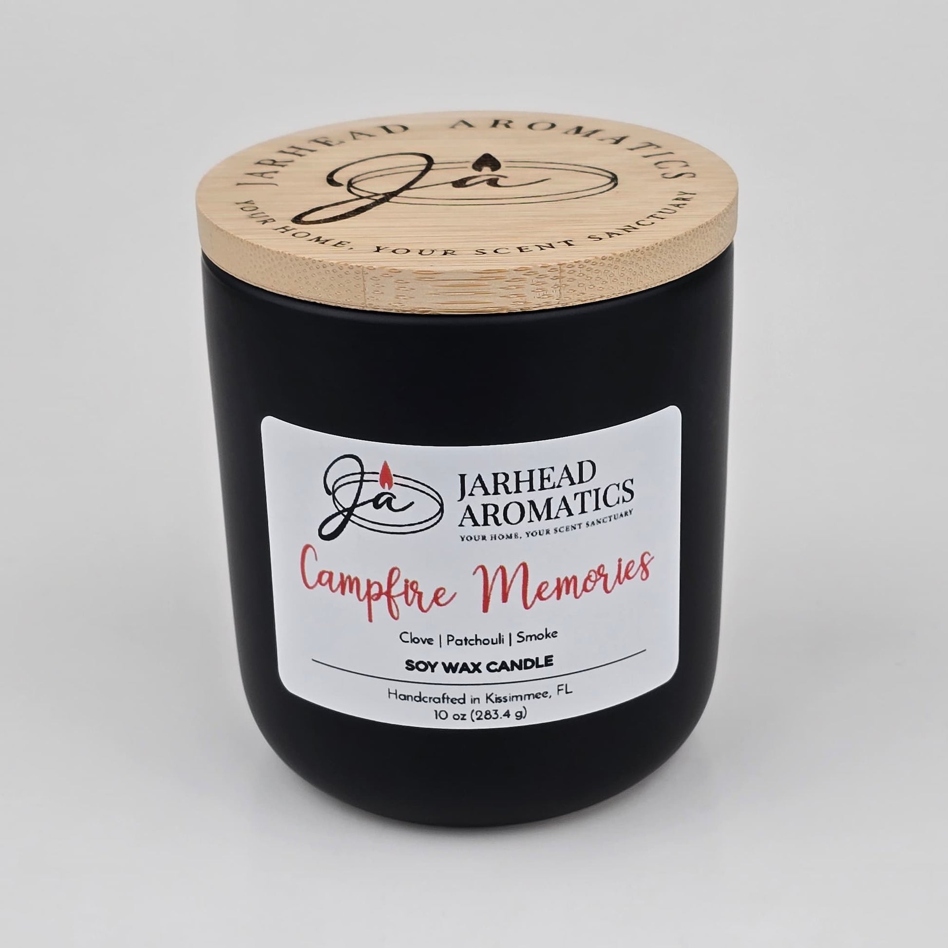 A black soy wax candle jar with a wooden lid from Jarhead Aromatics, labeled "Campfire Memories," features notes of smoky embers, maple, moss, and patchouli. This handcrafted autumn candle weighs 10 oz (283.4g) and is made in Kissimmee, FL.