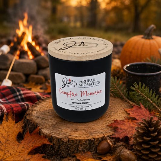 A Jarhead Aromatics Campfire Memories Soy Wax Candle—with smoky embers, maple, moss, and patchouli notes—rests on a log near autumn leaves. In the background, a campfire flickers beside a mug, pinecone, and cozy blanket, evoking a warm and rustic outdoor setting. This fall candle fills the air with nostalgic fragrances reminiscent of evenings spent by the fire.