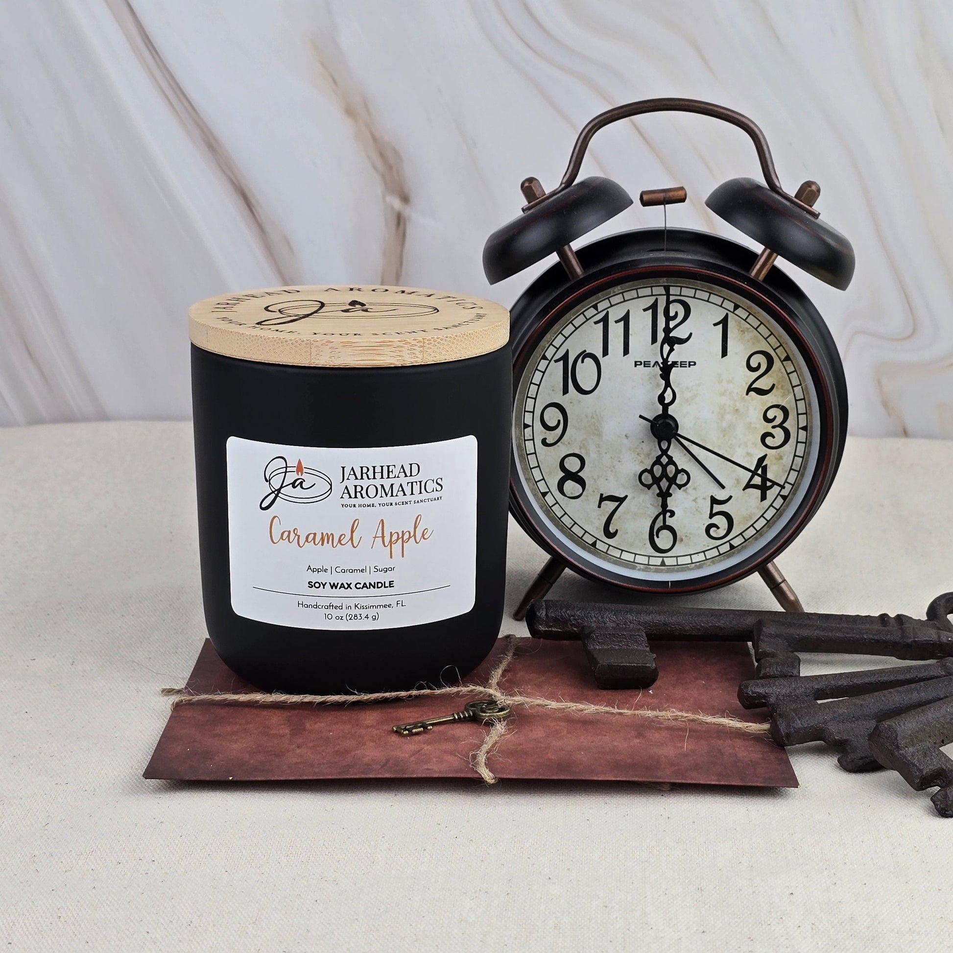 A Jarhead Aromatics Caramel Apple - Soy Wax Candle in black sits beside a vintage black alarm clock and a decorative key atop a brown envelope, all set on light marble—evoking the classic charm of caramel apples.