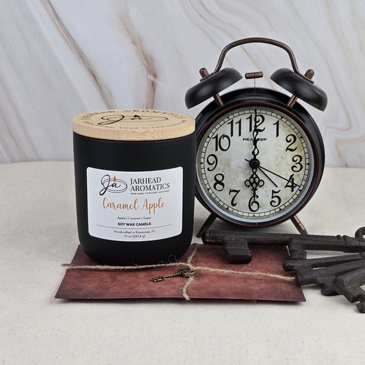 A Jarhead Aromatics Caramel Apple - Soy Wax Candle in black sits beside a vintage black alarm clock and a decorative key atop a brown envelope, all set on light marble—evoking the classic charm of caramel apples.