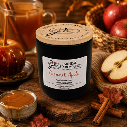 A Jarhead Aromatics Caramel Apple Soy Wax Candle rests on a wooden table surrounded by caramel, cinnamon sticks, apples, and a caramel apple, filling the space with its warm apple cider, caramel, and sugar fragrance for a cozy autumn ambiance.
