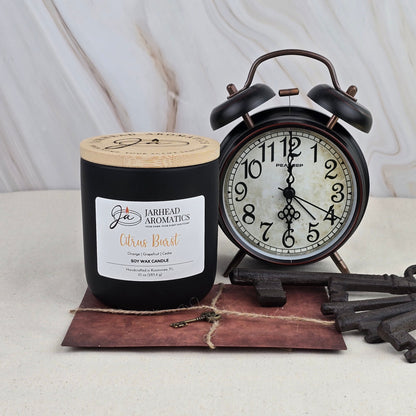 A black Citrus Burst - Soy Wax Candle by Jarhead Aromatics sits beside a vintage black alarm clock on a brown envelope with a key, set against a light marbled background.