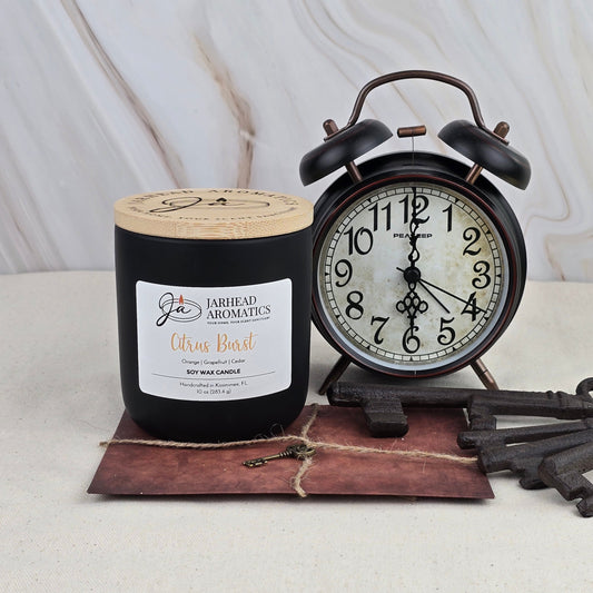 A black Citrus Burst - Soy Wax Candle by Jarhead Aromatics sits beside a vintage black alarm clock on a brown envelope with a key, set against a light marbled background.