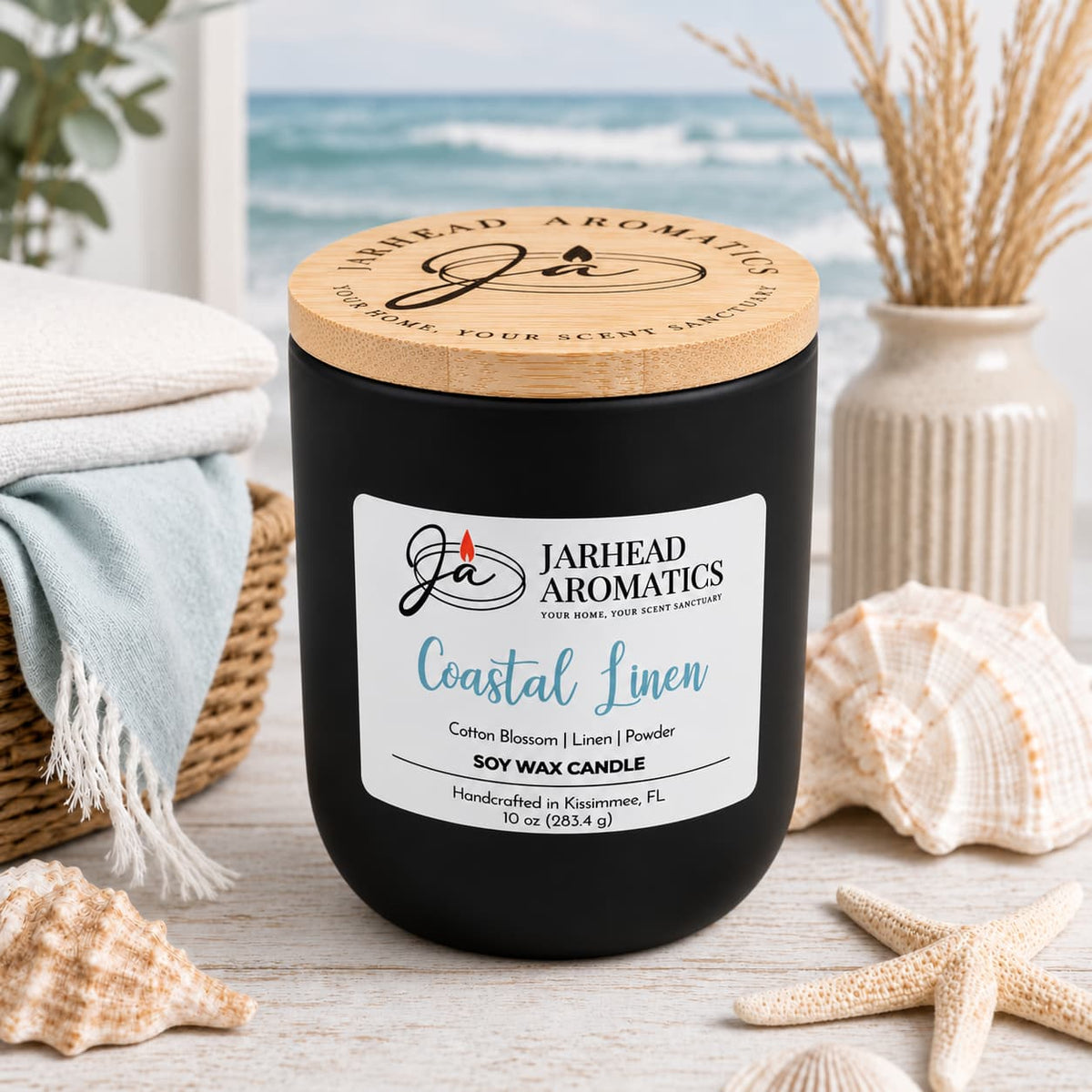 A black Coastal Linen Soy Wax Candle by Jarhead Aromatics with a wooden lid sits on a wooden surface surrounded by seashells and folded towels, with an ocean view in the background.