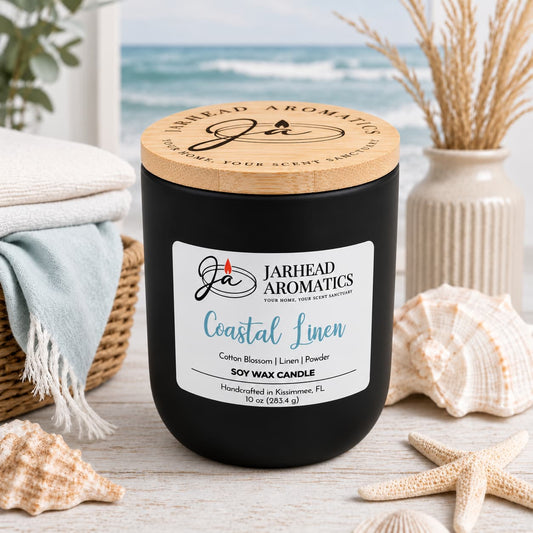 A black Coastal Linen Soy Wax Candle by Jarhead Aromatics with a wooden lid sits on a wooden surface surrounded by seashells and folded towels, with an ocean view in the background.