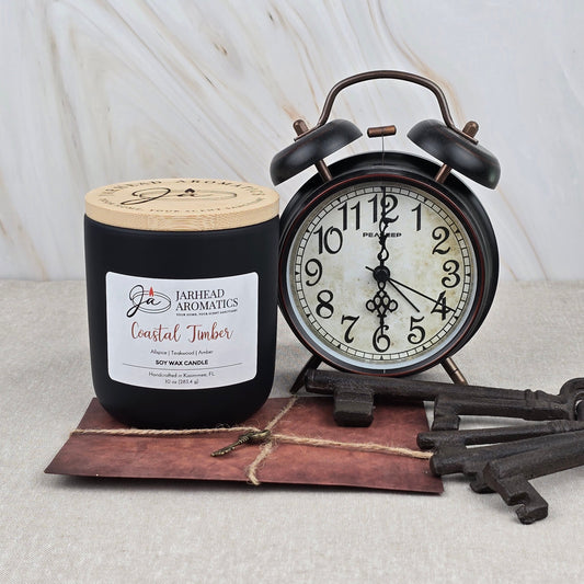 A Coastal Timber Soy Wax Candle | Teakwood, Leather & Amber by Jarhead Aromatics sits beside a vintage black alarm clock on a brown notepad with metal keys tied with twine, arranged on a light surface—this masculine candle elevates any space.