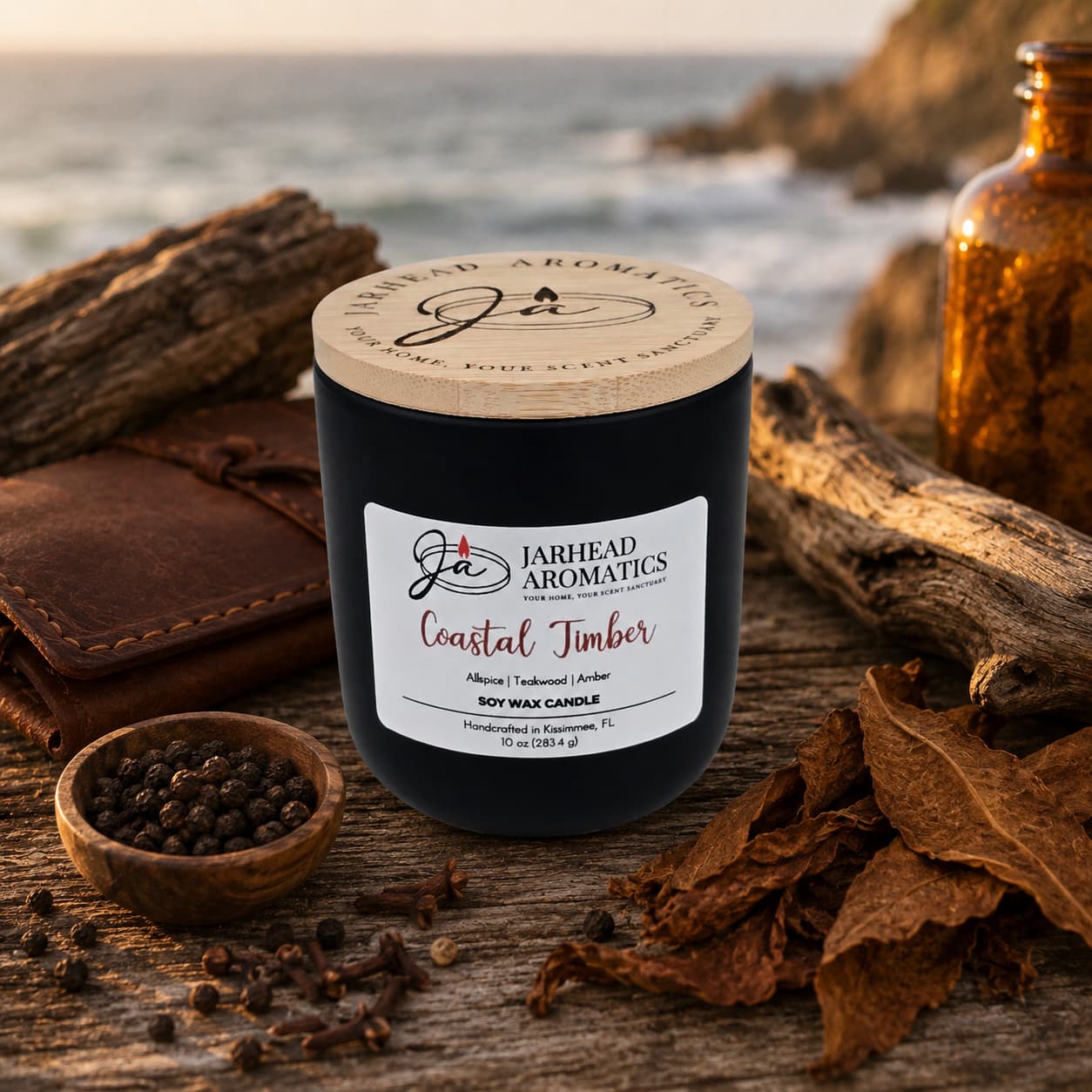 A Jarhead Aromatics Coastal Timber Soy Wax Candle | Teakwood, Leather & Amber Candle rests on driftwood by the sea, encircled by dried tobacco leaves, peppercorns, a notebook, and twine. This masculine candle fills the air with a distinctive blend of teakwood, leather, and amber—evoking the rugged character of coastal landscapes.