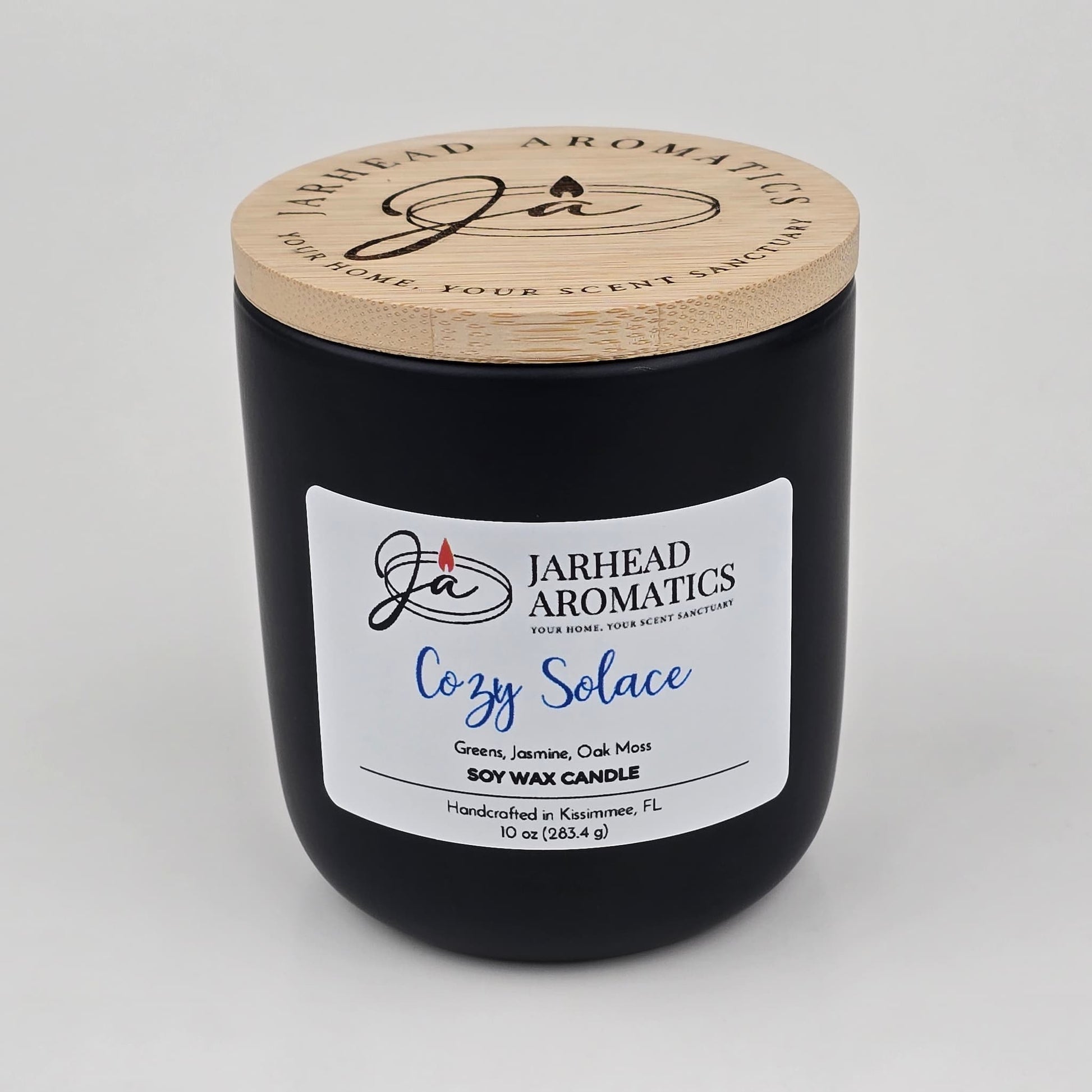 A black soy wax candle jar with a wooden lid from Jarhead Aromatics, the Cozy Solace Soy Wax Candle showcases a blend of fresh greens, jasmine, oak moss, and musk. Hand-poured in Kissimmee, FL, this autumn-inspired candle captures an inviting seasonal fragrance.