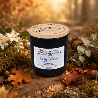 A hand-poured Cozy Solace Soy Wax Candle from Jarhead Aromatics rests atop autumn leaves and moss, surrounded by flowers and foliage, with a blurred forest backdrop—capturing the essence of fresh greens, jasmine, oak moss, and musk in a warm fall fragrance.