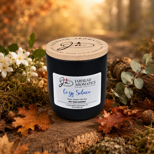 A hand-poured Cozy Solace Soy Wax Candle from Jarhead Aromatics rests atop autumn leaves and moss, surrounded by flowers and foliage, with a blurred forest backdrop—capturing the essence of fresh greens, jasmine, oak moss, and musk in a warm fall fragrance.