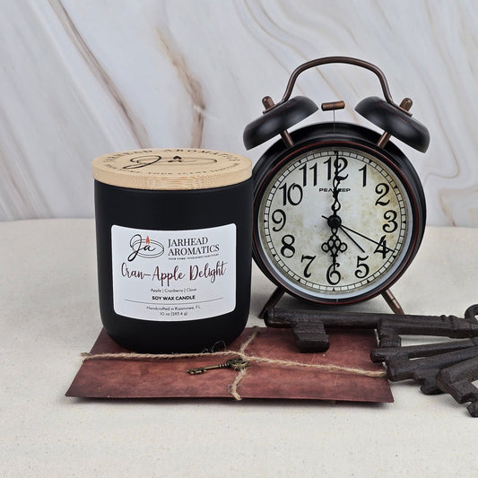 A black jar of Cran-Apple Delight - Soy Wax Candle by Jarhead Aromatics sits beside a vintage-style black alarm clock on a brown notepad tied with twine, all set against a light marbled background.