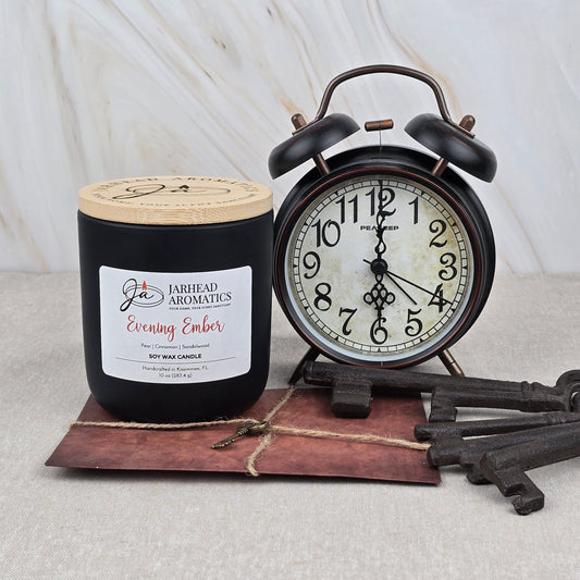 An Evening Ember Soy Wax Candle | Plum, Sandalwood & Smoke by Jarhead Aromatics rests next to a vintage black alarm clock, three antique keys, and a tied brown envelope on marble.