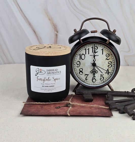A black Fairytale Spice - Soy Wax Candle by Jarhead Aromatics rests next to a vintage black alarm clock, an old key, and a brown envelope on a beige surface against a marble backdrop.