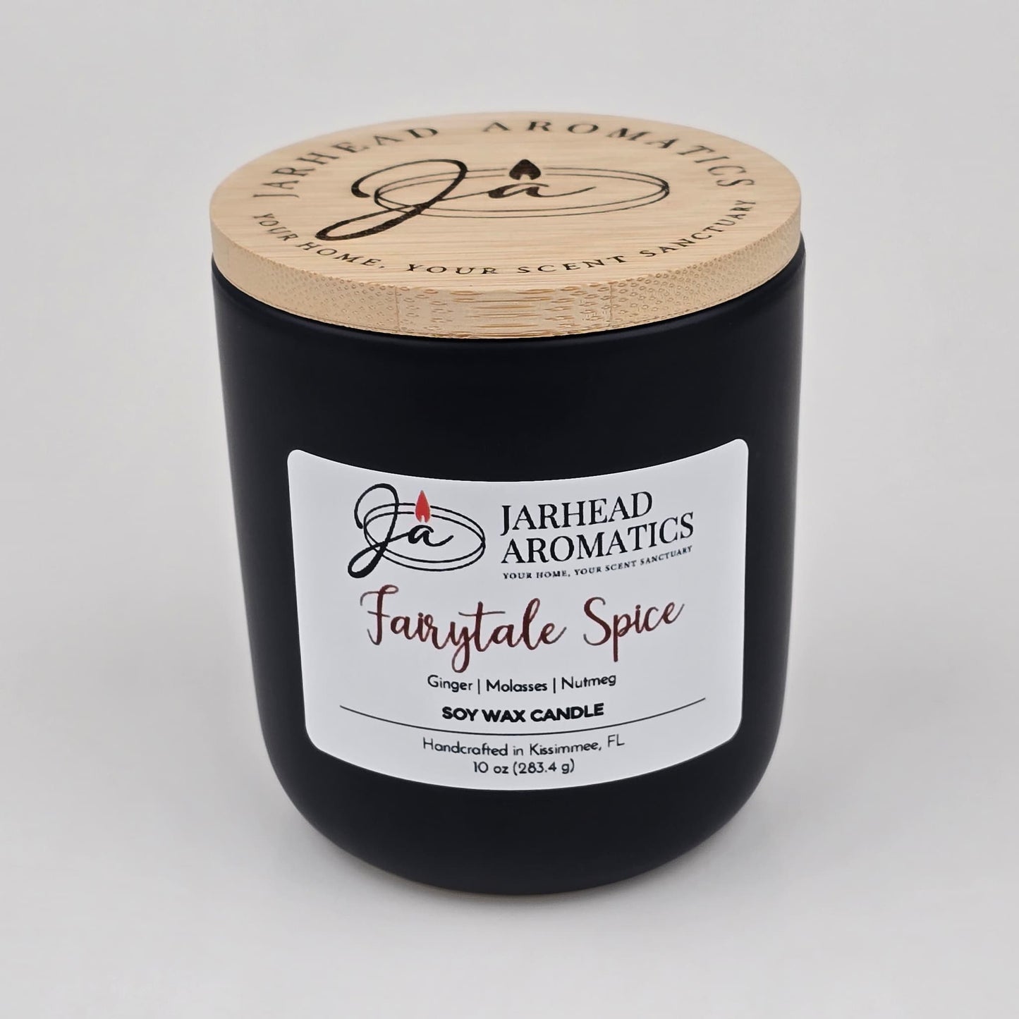 A black candle jar with a wooden lid from Jarhead Aromatics, labeled Fairytale Spice Soy Wax Candle | Gingerbread, Cinnamon & Vanilla Candle. This handcrafted holiday candle is made in Kissimmee, FL with soy wax and features festive scents of gingerbread, cinnamon, and vanilla—perfect for bringing warmth and cheer to your home.