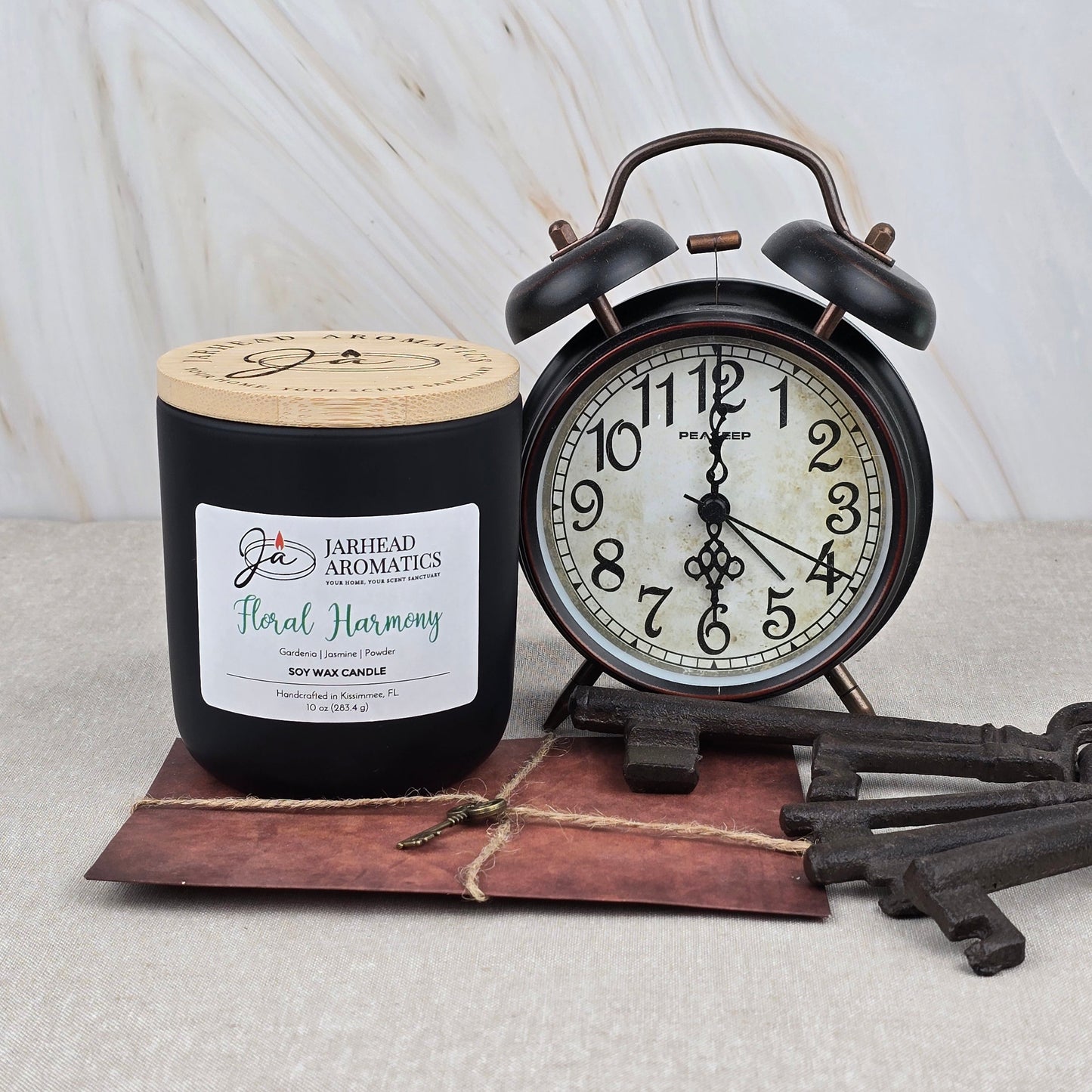 A Floral Harmony Soy Wax Candle by Jarhead Aromatics stands beside a vintage black alarm clock set to 2:00, both atop a string-tied brown envelope with two antique keys, all placed on a light marble surface.