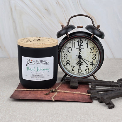 A Floral Harmony Soy Wax Candle by Jarhead Aromatics stands beside a vintage black alarm clock set to 2:00, both atop a string-tied brown envelope with two antique keys, all placed on a light marble surface.