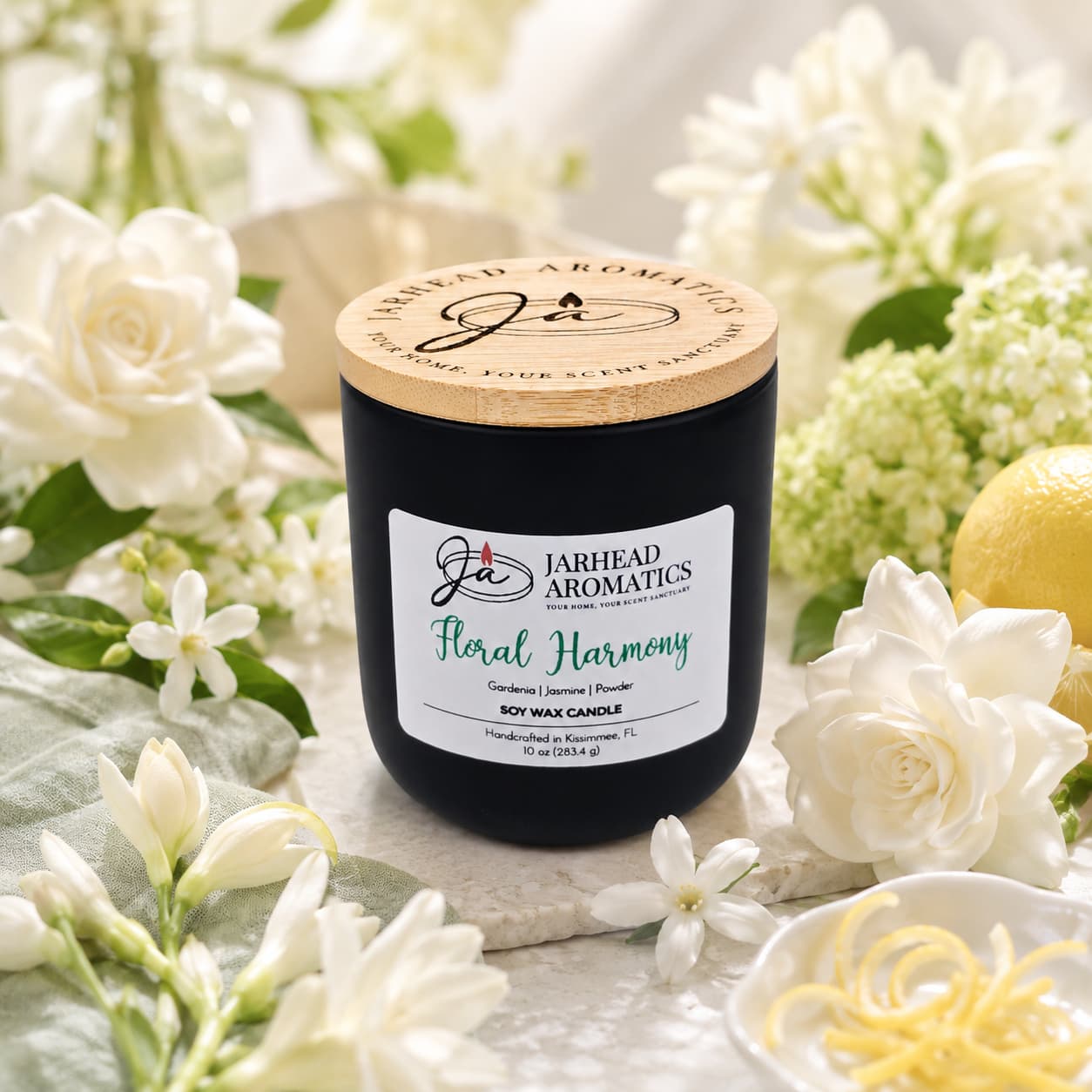 A matte black candle jar with a wooden lid, labeled "Floral Harmony Soy Wax Candle | Gardenia, Jasmine & Tuberose Candle" by Jarhead Aromatics, is surrounded by white blossoms, fresh green leaves, and a vibrant lemon with twisted zest on a softly lit surface. This hand-poured soy wax candle fills your space with the long-lasting fragrance of gardenia, jasmine, and tuberose—bringing an essence of floral harmony to any room.