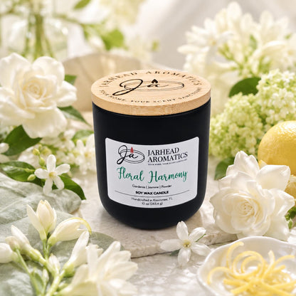 A matte black candle jar with a wooden lid, labeled "Floral Harmony Soy Wax Candle | Gardenia, Jasmine & Tuberose Candle" by Jarhead Aromatics, is surrounded by white blossoms, fresh green leaves, and a vibrant lemon with twisted zest on a softly lit surface. This hand-poured soy wax candle fills your space with the long-lasting fragrance of gardenia, jasmine, and tuberose—bringing an essence of floral harmony to any room.