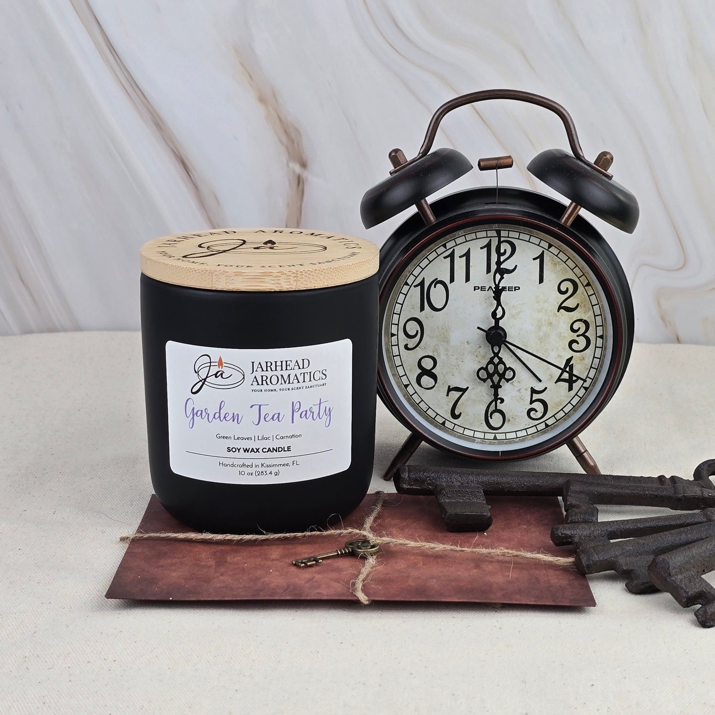A Jarhead Aromatics Garden Tea Party Soy Wax Candle sits beside a vintage black alarm clock showing 9:16, with old keys and a brown string-tied envelope in the foreground, against a light marble background.