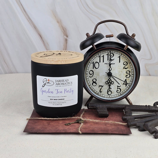A Jarhead Aromatics Garden Tea Party Soy Wax Candle sits beside a vintage black alarm clock showing 9:16, with old keys and a brown string-tied envelope in the foreground, against a light marble background.