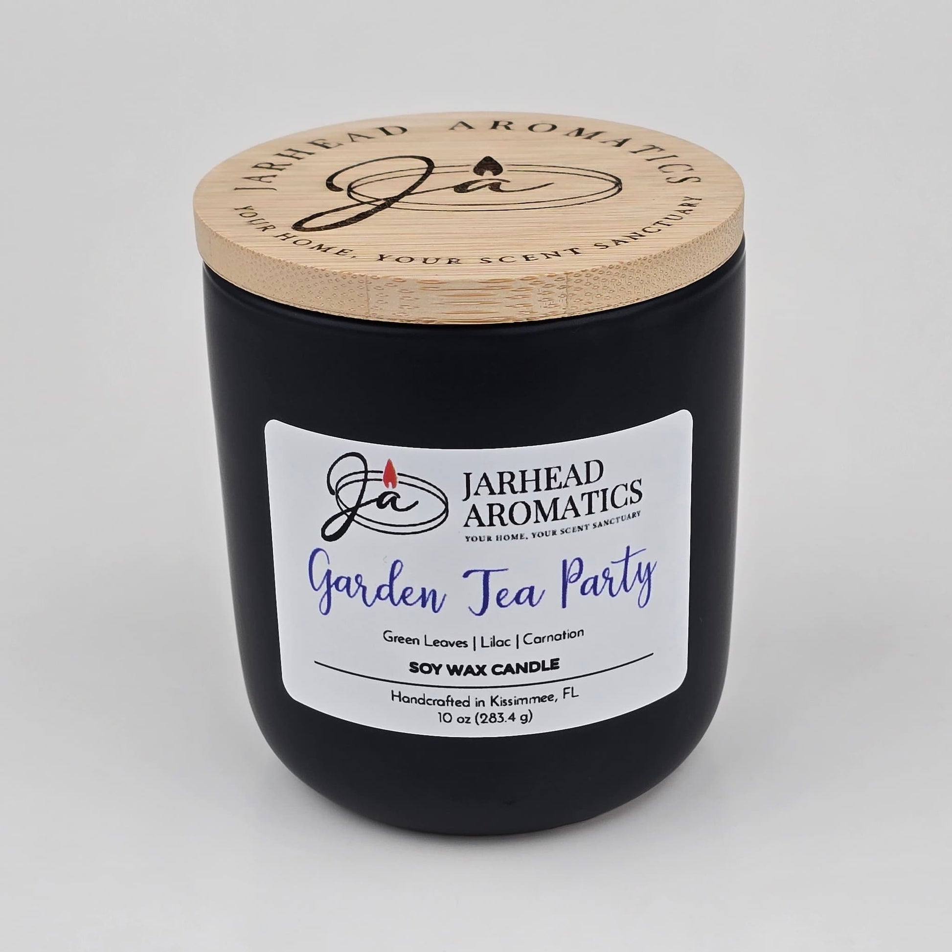 A black Jarhead Aromatics hand-poured soy wax candle, labeled Garden Tea Party, featuring notes of rose, jasmine, and green floral accords. This candle is handcrafted in Kissimmee, FL and comes with a wooden lid.