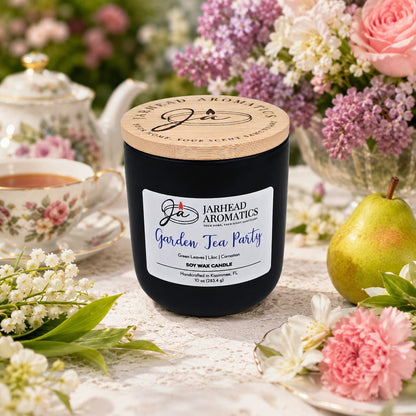A Garden Tea Party Soy Wax Candle by Jarhead Aromatics sits on a table adorned with flowers, a pear, and a teacup, creating an elegant, garden-inspired setting. This rose, jasmine & green floral candle fills the air with delicate aromas, perfectly complementing the charming scene.