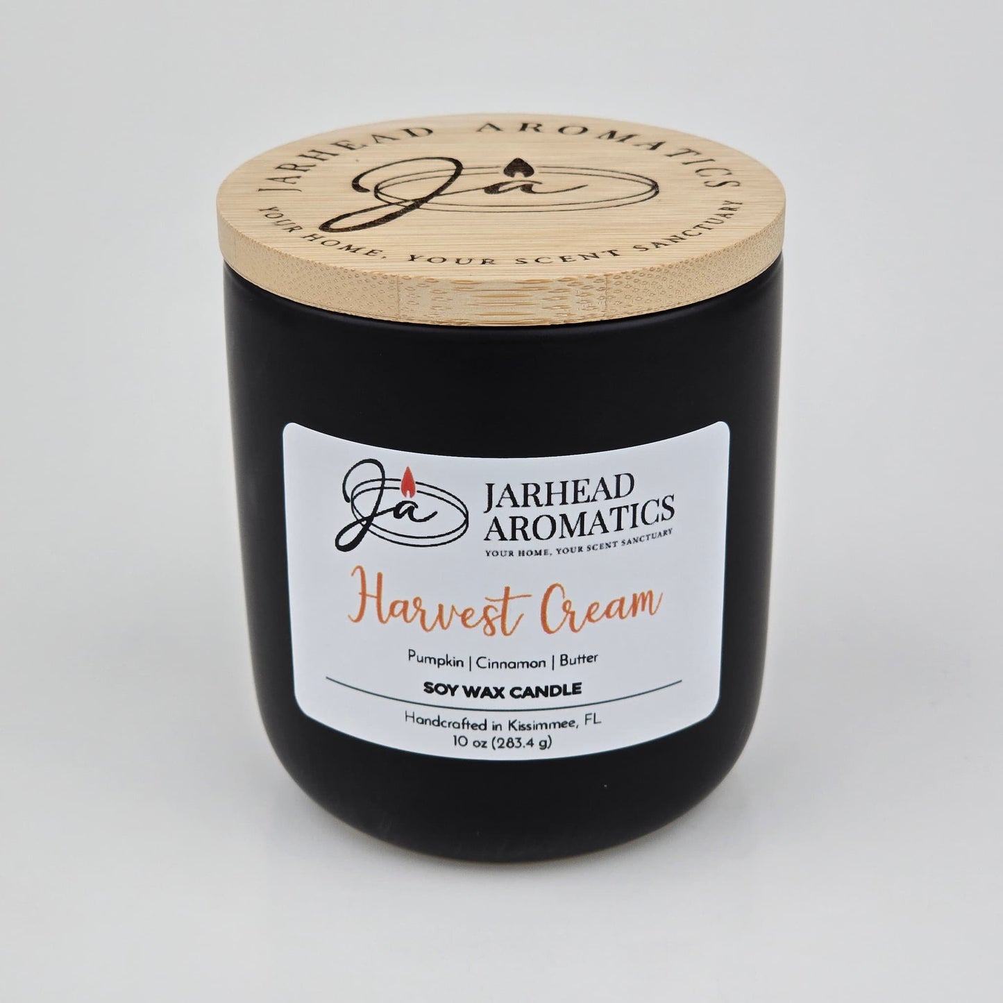 A black candle jar with a wooden lid from Jarhead Aromatics, labeled Harvest Cream Soy Wax Candle, features inviting notes of pumpkin spice, brown sugar, and vanilla. This handcrafted fall candle is made in Kissimmee, FL and fills your space with a warm, cozy autumn fragrance.