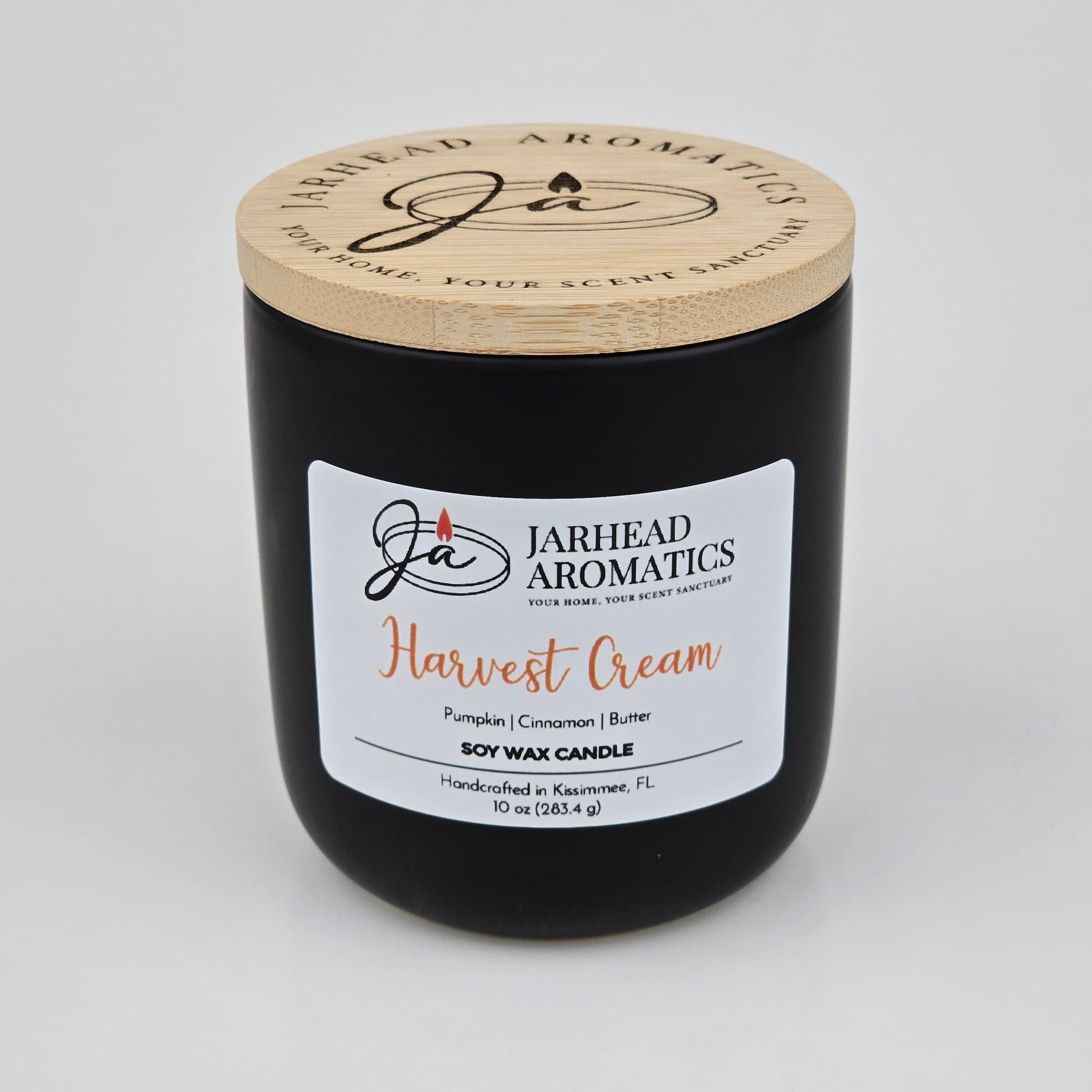 A black candle jar with a wooden lid from Jarhead Aromatics, labeled Harvest Cream Soy Wax Candle, features inviting notes of pumpkin spice, brown sugar, and vanilla. This handcrafted fall candle is made in Kissimmee, FL and fills your space with a warm, cozy autumn fragrance.