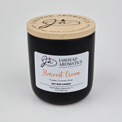 A black candle jar with a wooden lid from Jarhead Aromatics, labeled Harvest Cream Soy Wax Candle, features inviting notes of pumpkin spice, brown sugar, and vanilla. This handcrafted fall candle is made in Kissimmee, FL and fills your space with a warm, cozy autumn fragrance.