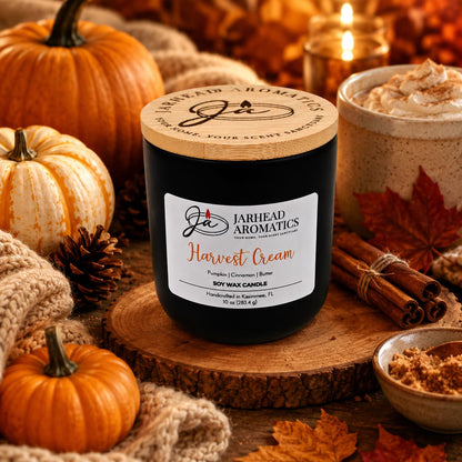 A Jarhead Aromatics Harvest Cream Soy Wax Candle—infused with notes of pumpkin spice, brown sugar, and vanilla—rests on a cozy table surrounded by pumpkins, pinecones, cinnamon sticks, a warm drink, and knit blankets, filling the air with an inviting fall fragrance and creating a festive autumn atmosphere.