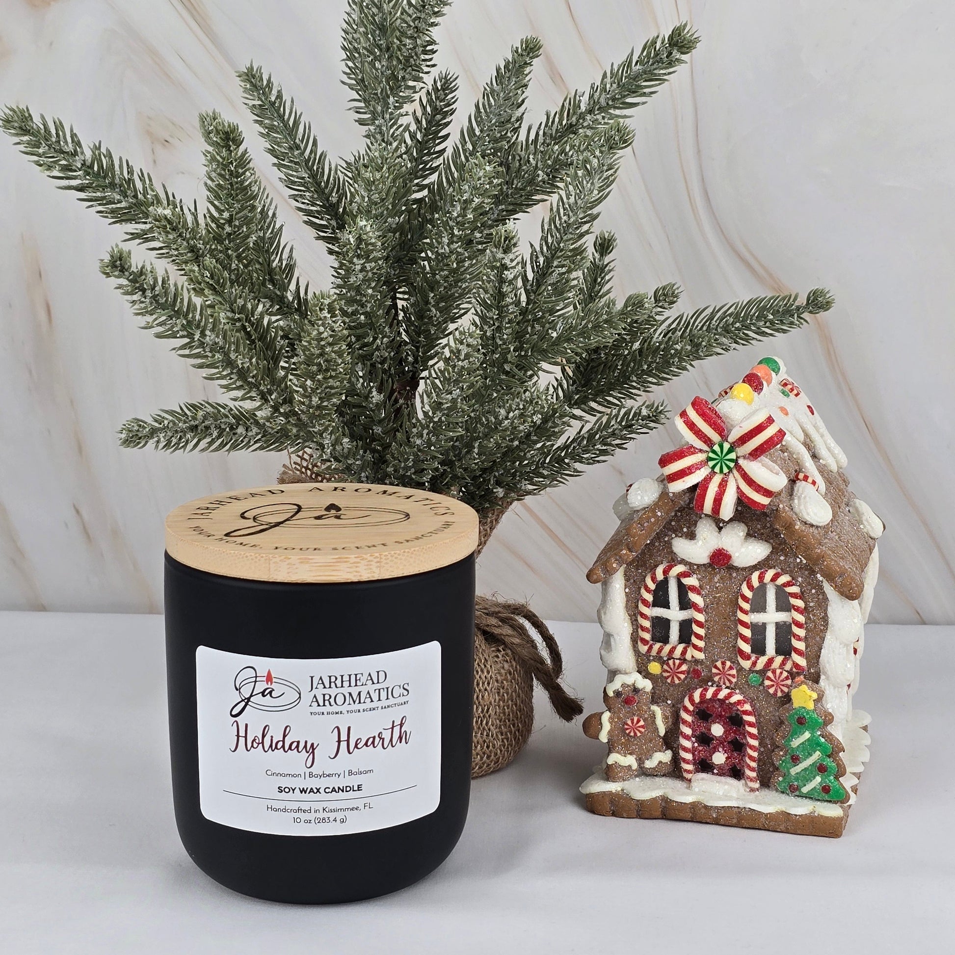 The Jarhead Aromatics Holiday Hearth - Soy Wax Candle from the Winter Collection sits beside a faux pine tree and decorated gingerbread house, all set on a marble background for a festive holiday setting.