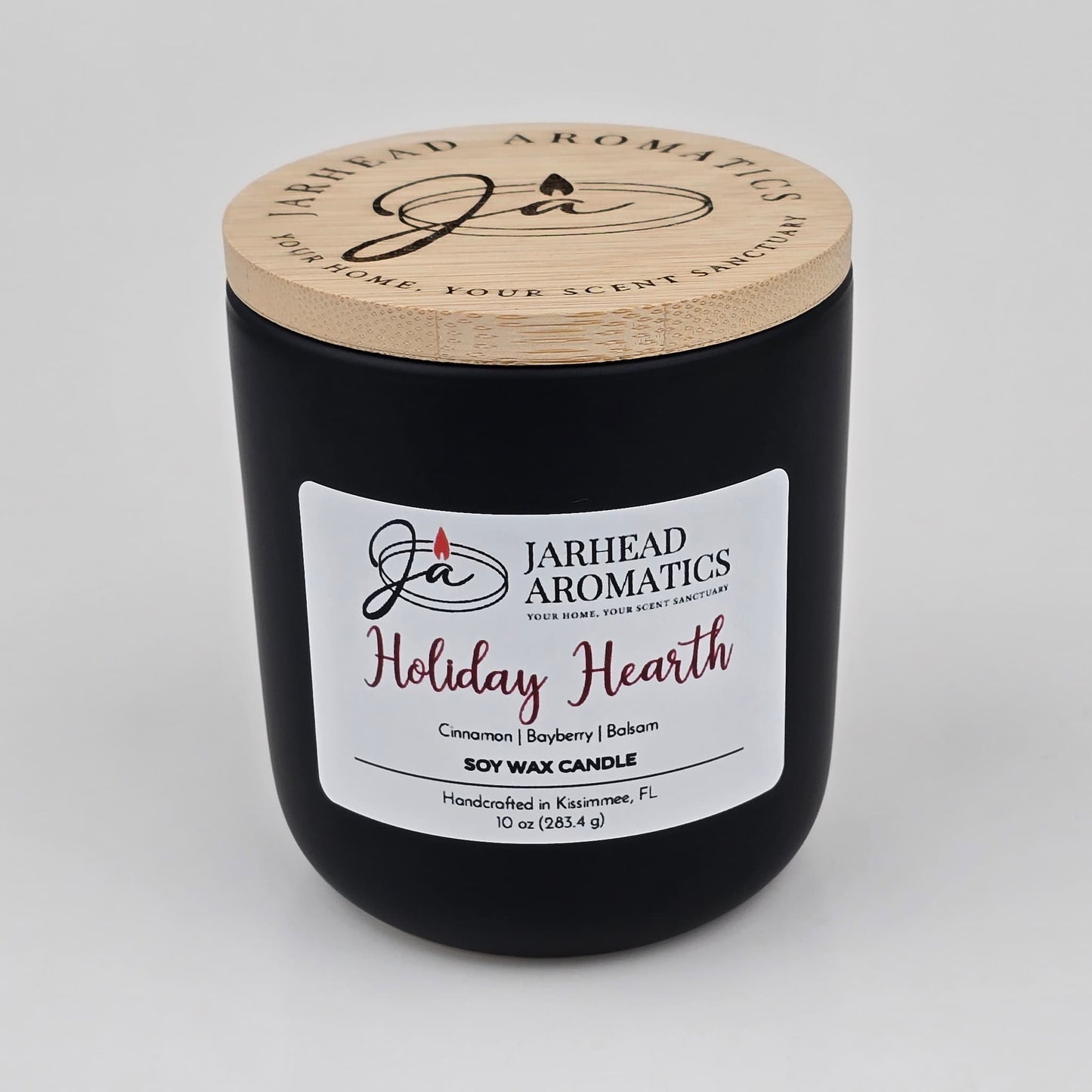 A black candle jar with a wooden lid, labeled Jarhead Aromatics, contains the Holiday Hearth Soy Wax Candle—handcrafted in Kissimmee, FL. This holiday candle features warm scents of cinnamon clove, bayberry, and balsam.