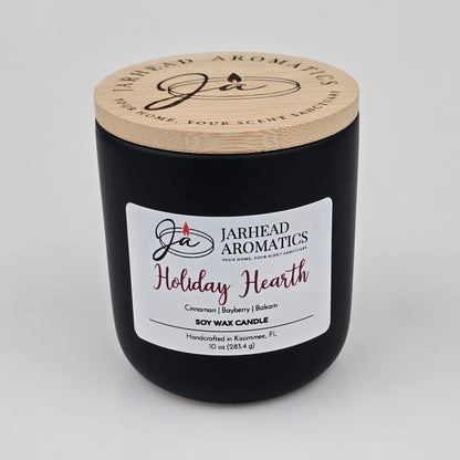 A black candle jar with a wooden lid, labeled Jarhead Aromatics, contains the Holiday Hearth Soy Wax Candle—handcrafted in Kissimmee, FL. This holiday candle features warm scents of cinnamon clove, bayberry, and balsam.