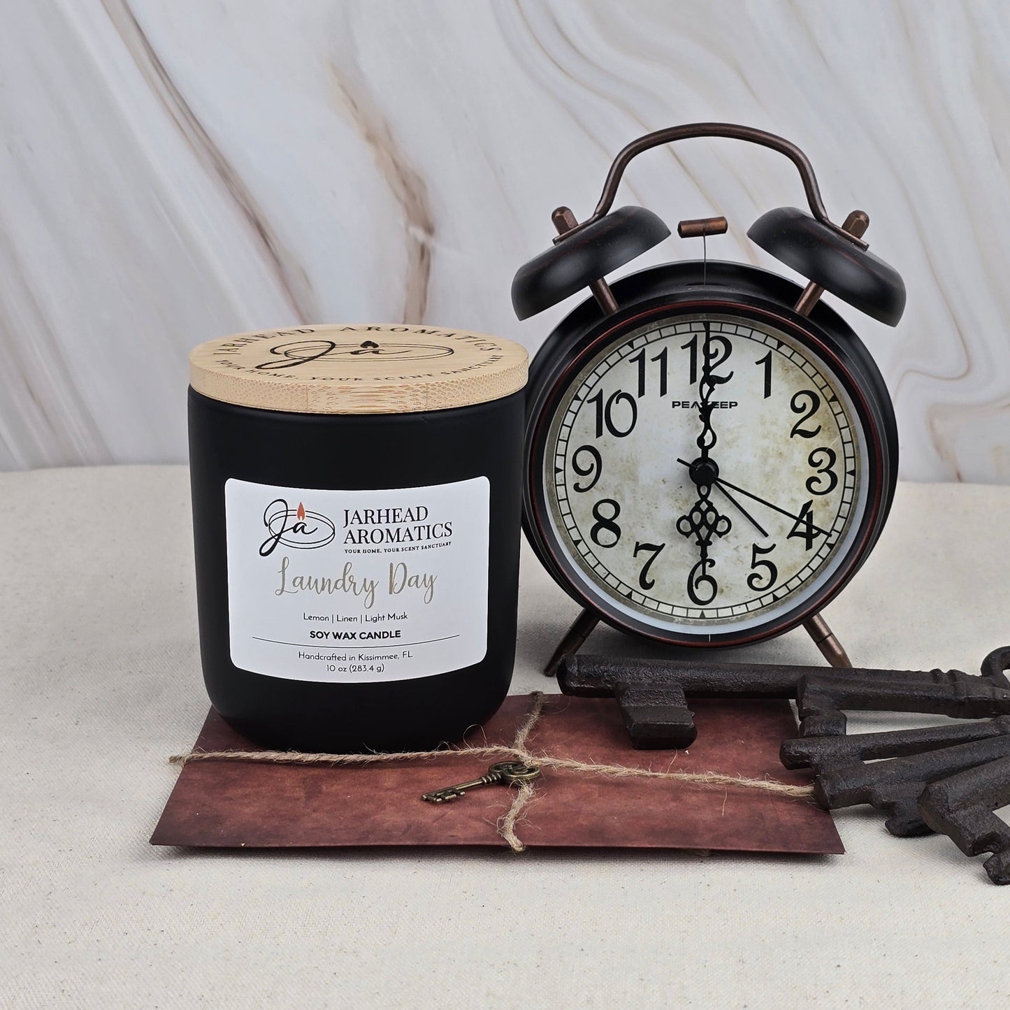 A black Jarhead Aromatics Laundry Day soy wax candle with a sandalwood scent sits beside a vintage black alarm clock showing 8:00 on a small brown envelope with large antique keys, all set against a light marble background.