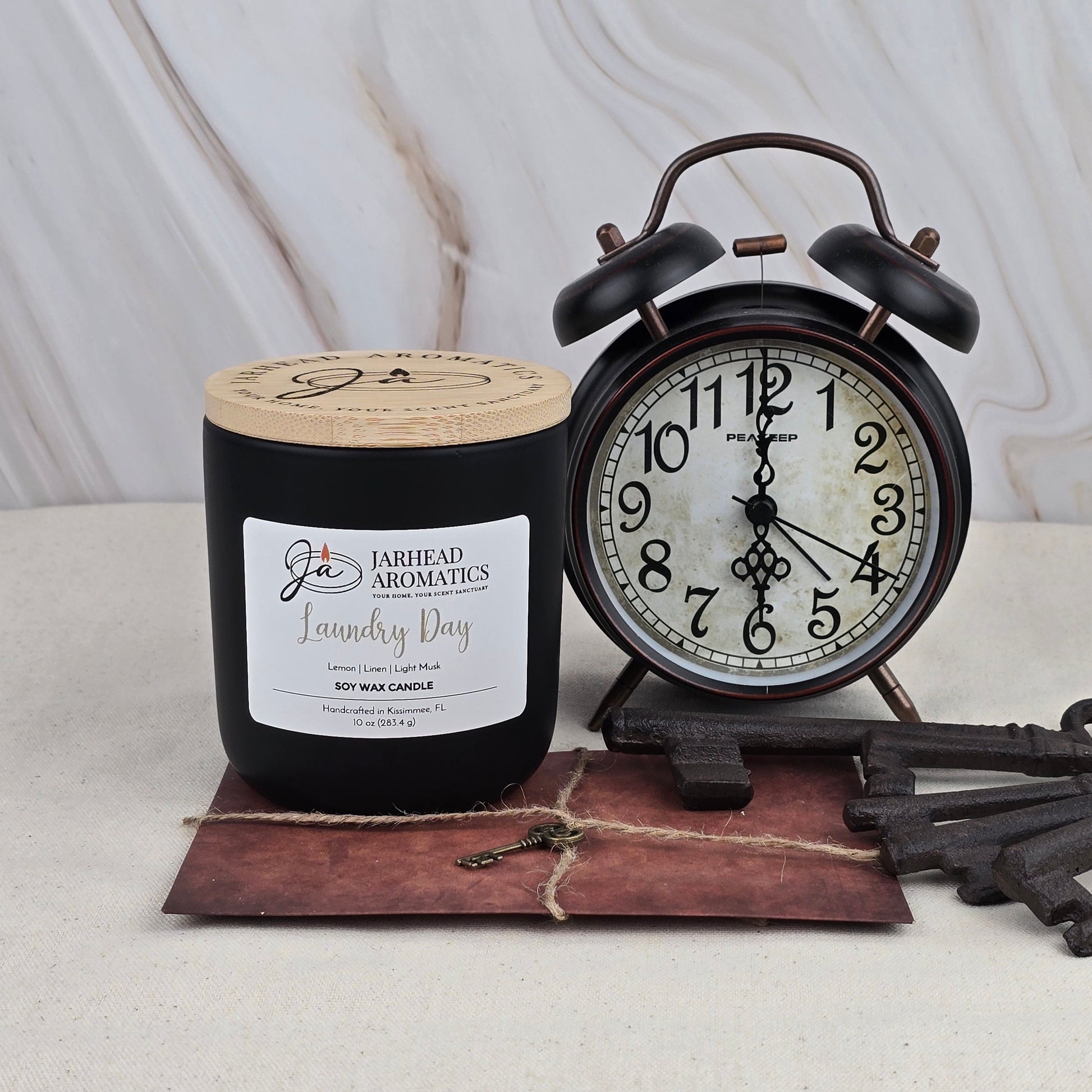 A black Jarhead Aromatics Laundry Day soy wax candle with a sandalwood scent sits beside a vintage black alarm clock showing 8:00 on a small brown envelope with large antique keys, all set against a light marble background.