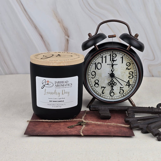 A black Jarhead Aromatics Laundry Day soy wax candle with a sandalwood scent sits beside a vintage black alarm clock showing 8:00 on a small brown envelope with large antique keys, all set against a light marble background.