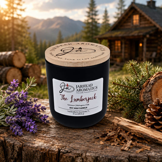 A black Jarhead Aromatics The Lumberjack Soy Wax Candle (Cedar, Oak & Mahogany) sits on a rustic wooden surface outdoors amid lavender, pine branches, and logs with a log cabin and mountains behind, exuding a masculine woodland cabin scent.