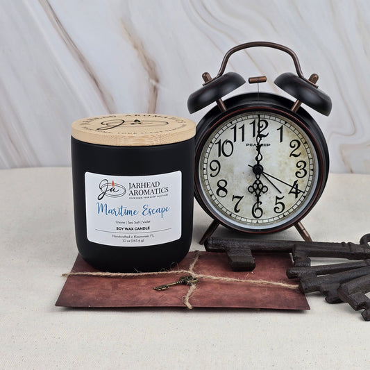 A black Maritime Escape - Soy Wax Candle by Jarhead Aromatics rests on a rustic envelope beside a vintage black twin-bell alarm clock showing 11:21, set against a light marbled background.