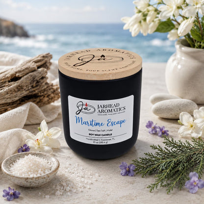A black Jarhead Aromatics Maritime Escape Soy Wax Candle, featuring a wooden lid engraved with the brand name, rests on a sandy surface surrounded by flowers and driftwood, with sea salt and ocean views in the background. The coastal candle blends notes of sea salt, ozone, and cedar.