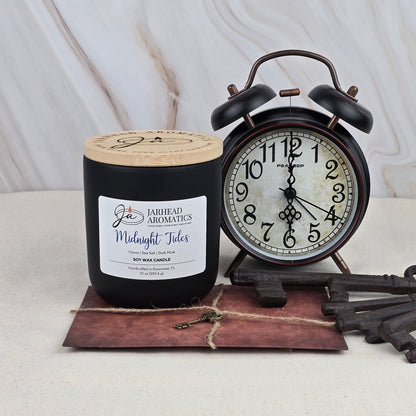 A Jarhead Aromatics Midnight Tides Soy Wax Candle in a black jar, featuring a clean ozonic scent from essential oils, is displayed beside a vintage black alarm clock at 7:05, with twine-tied envelope and antique keys on a beige surface.