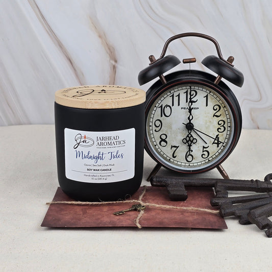 A Jarhead Aromatics Midnight Tides Soy Wax Candle in a black jar, featuring a clean ozonic scent from essential oils, is displayed beside a vintage black alarm clock at 7:05, with twine-tied envelope and antique keys on a beige surface.