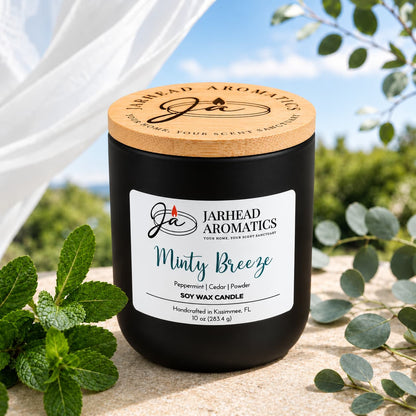 A black Minty Breeze Soy Wax Candle with a wooden lid sits on a stone surface next to fresh mint sprigs.