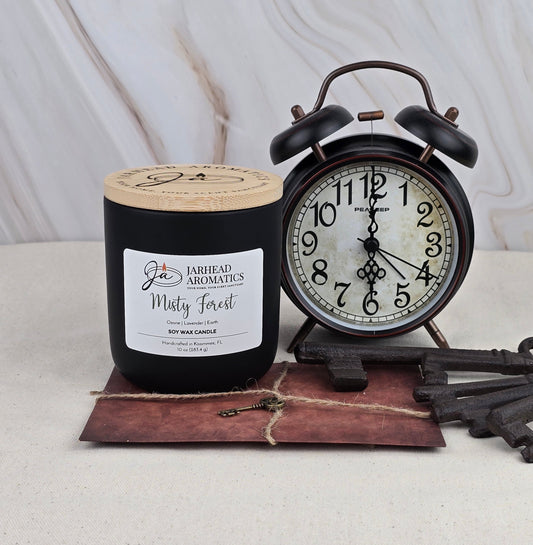 A black Jarhead Aromatics Misty Forest - Soy Wax Candle with woodsy scents sits on a brown envelope next to a vintage black alarm clock and two old keys, all arranged on a light marbled surface.
